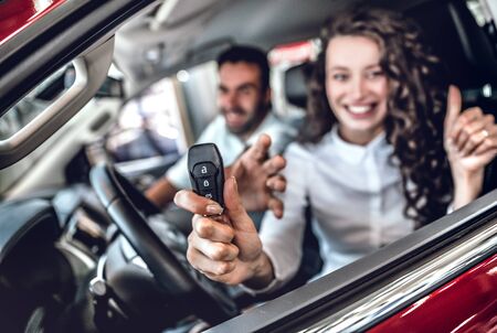 Close up of attractive woman showing car key through the window. Nice young couple sitting inside the new car at the dealership. Focus on keyの写真素材