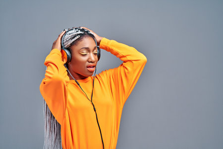 Cheerful afro american woman with closed eyes listening music in headphones over gray background....