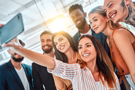 Multi-ethnic group of adult people smiling at camera while taking selfie photo in office. Sunlight background. Selective focusの写真素材