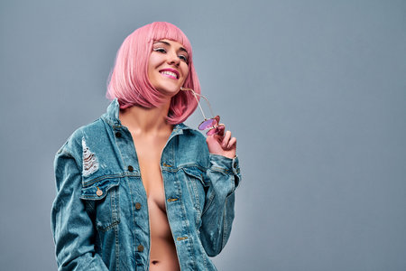 Young attractive stylish woman wearing blue jeans jacket and holding pink sunglasses on studio background. Close up view portrait, trendy lifestyle. Copy spaceの写真素材