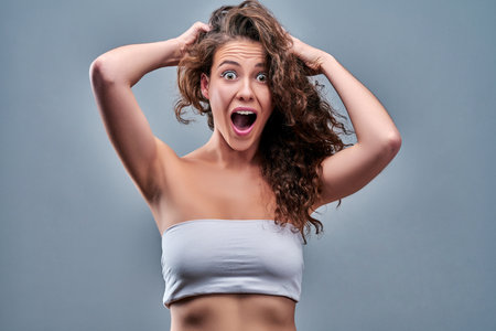 Photo of curly wavy brunette horrified feared girl holding her head with face expressing disappointment isolated over gray background.の写真素材