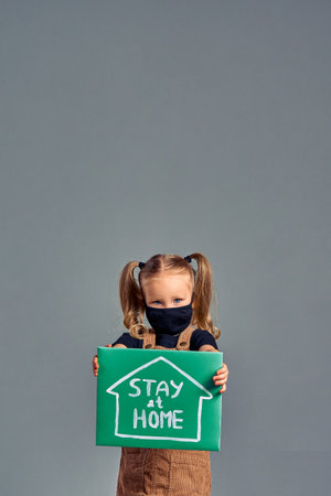Little girl in medical mask holding in hands poster with Stay at home word. Coronavirus concept. Isolated gray. Vertical photoの写真素材