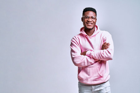 Handsome african american young man in glasses and pink hoodie standing with arms crossed on gray background. Copy spaceの写真素材
