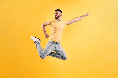 A young man dressed in a yellow T-shirt and light jeans jumps against a yellow wall, straightening...