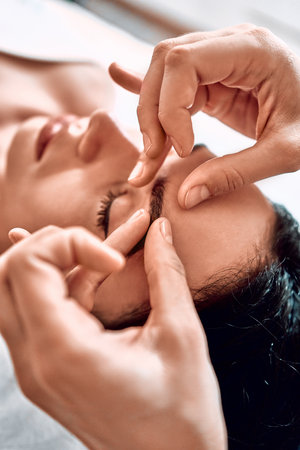 The process of sculpting facial massage in the cosmetologist s office. Beautiful woman looks after her appearance. Beautician massages her client, removes signs of aging from the face. Vertical photo. Close up viewの写真素材