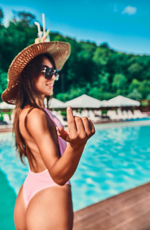 Follow me, young laughing girl asking you to follow her, waving hand. Woman in pink body swimsuit, on hot sunny day. Summer holiday at luxury hotel, background swimming pool. selective focusの写真素材