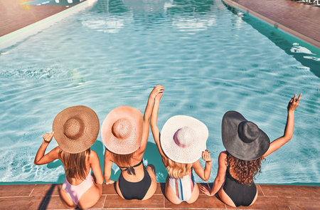 Four beautiful young woman with sun hat sitting by the poolside of a resort swimming pool during...