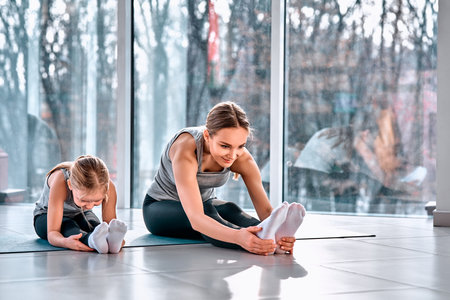 Side view shot of young smiling sporty woman and her little daughter sitting on yoga mat, doing warm up exercise before training. Family preparing to workout. Wellbeing, wellness conceptの写真素材