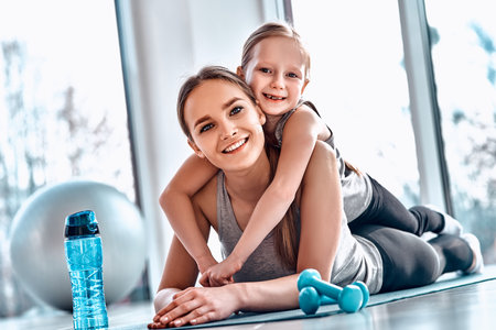 A little girl lies on her mother's back while resting after workout in gym atmosphere with dumbbells, mat, fitness ball, bottle of water on the floor. Healthy family leisure conceptの写真素材