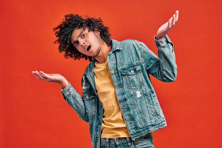 Nervous young man shrugging hands on isolated orange background. Confused eastern curly student...