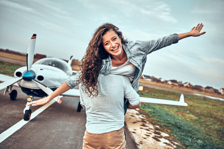 High flying romance. Front view of smiling young woman piggybacking on her boyfriend while keeping arms outstretched. Private plane backgroundの写真素材
