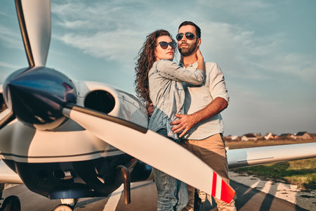 Beautiful and happy young couple posing in front of an old retro private propeler airplane and representing happines wealt and rich life conceptの写真素材