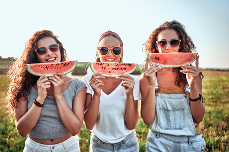 Three nice girls having fun eating watermelon In the park. Excellent sunny weather. Beautiful figures. Super mood. Summer concept.の写真素材