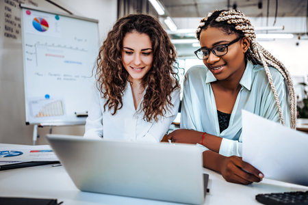 Two young beautiful women sit at desk looking laptop in modern office.の写真素材