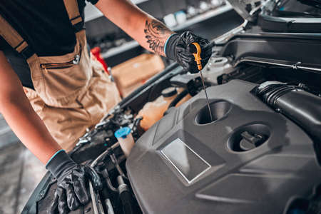 Concept of male car mechanic holding dipstick checking level oil on the car engine, for repair and maintenance services, wearing a black glove and beige garment overalls with front car engine. Close up view. Cropped image.の写真素材