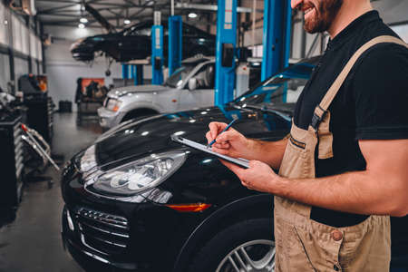 Cropped image of handsome mechanic in uniform making notes while standing in auto service. Side view.の写真素材