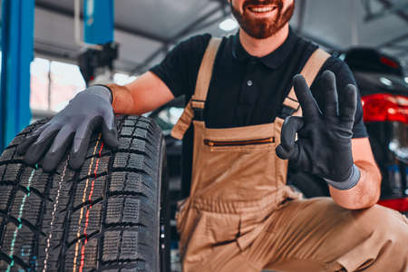 Trustworthy mechanic showing ok gesture, holding car tire and smiling on the car repair shop. Close up view. Cropped image.の写真素材