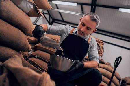 Man pouring coffee from a bag into a bowl for tasting, against the background of a warehouse. Bottom...