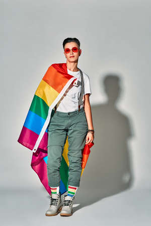 Happy beautiful woman in red eyewear smiling while posing with rainbow flag isolated over white...
