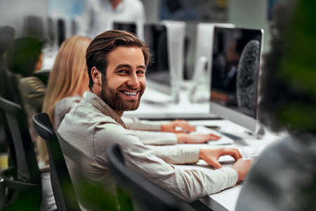 Handsome business man works at the computer with colleagues in the office and smiles looking at the...