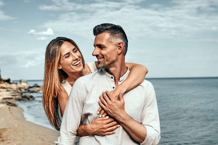 Woman holding her husband from behind standing at a beach on a sunny day. Romantic couple on standing near the sea together.の写真素材