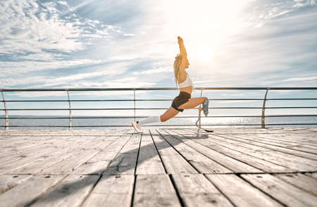 Sporty disabled athlete woman in sportswear with prosthetic leg standing in yoga pose on the bridge in front of the sea. Copy space. Full length. Side view.の写真素材