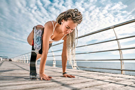 Young sportswoman with prosthesis doing exercise while working out outdoors and listening to music on earpods on a bridge near sea.の写真素材