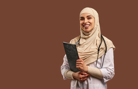 Portrait of a Muslim woman dressed in a white robe holding a folder and standing near a brown background. The concept of medicine, health care.Copy space.の写真素材