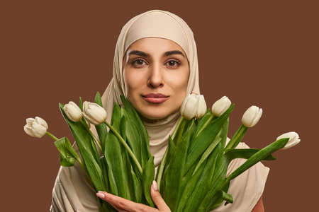 Close-up portrait of a beautiful Muslim woman dressed in a beige hijab who looks at the camera and holds a large bouquet of tulips on a brown background. beauty and facial care.の写真素材
