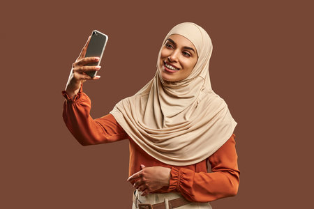 Beautiful Muslim woman in a beige hijab holds a phone in her hands, smiles and takes a selfie standing near a brown background.の写真素材