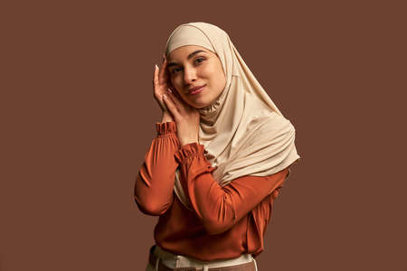 Modern, stylish, fashionable, beautiful Muslim woman dressed in a beige hijab, orange jacket and beige pants who looks at the camera and poses on a brown background.の写真素材
