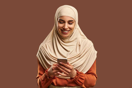 Beautiful Muslim woman in a beige hijab holds a phone in her hands and smiles. Advertising, technology, devices.の写真素材