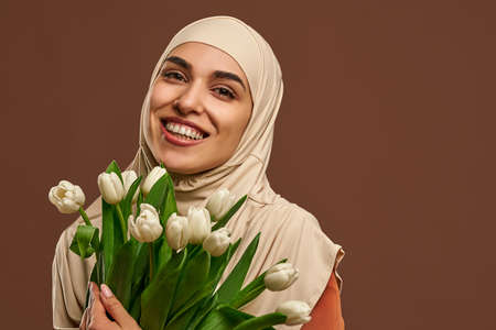 Close-up portrait of a beautiful muslim woman wearing a beige hijab smiling and looking at the camera and holding a big bouquet of tulips on a brown background.の写真素材
