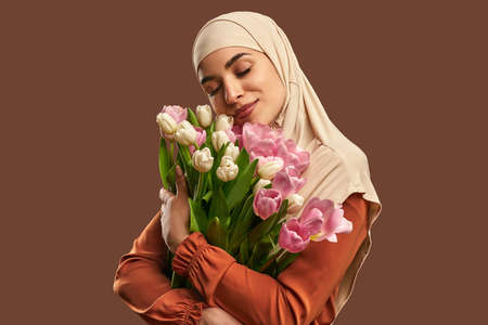Portrait of a beautiful Muslim woman wearing a beige hijab and holding a big bouquet of tulips on a brown background. The concept of female beauty, spring and femininity.の写真素材