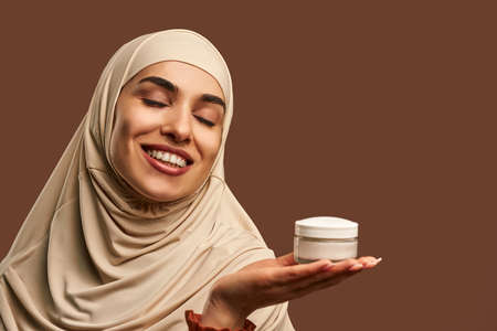 Portrait of beautiful modern muslim woman with natural make-up dressed in beige hijab smiling with closed eyes and holding cream in hand on brown background. Facial skin care, female beauty.の写真素材