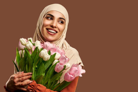 Beautiful Muslim woman dressed in a beige hijab smiles and looks away and holds a large bouquet of tulips on a brown background. copyspace.の写真素材