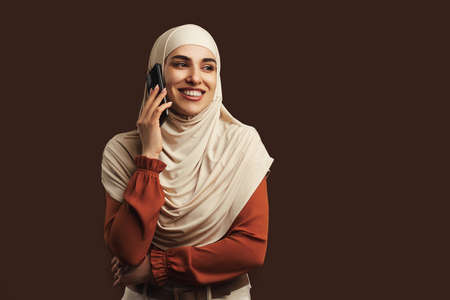 Beautiful muslim woman dressed in beige hijab holding phone smiling and talking on brown background. copyspace.の写真素材