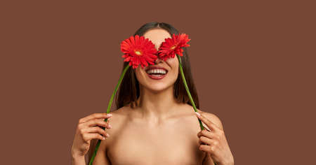 Portrait of a beautiful woman on a brown background holding two red flowers hiding their eyes and laughing. beauty concept.の写真素材