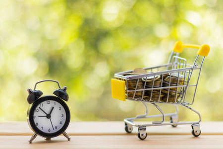 Alarm clock and mini shopping cart full of coins on wooden table, natural green blurred background. Business, finance, time, online shopping, money saving concept.の写真素材