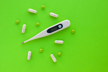Digital thermometer, green pills and white capsules isolated on green background. Epidemic,...