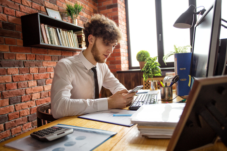Young business man looking at smartphone on working place at computerの写真素材