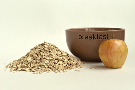 A handful of oat flakes, an apple and a brown cup for flakes with the inscription "breakfast" on a light background close-up.の写真素材