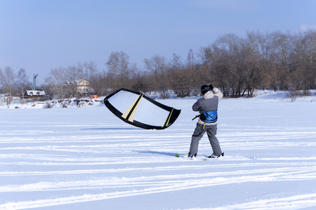 Snowkiter catches the wind with his kite, standing on the ice of a frozen pond against the shore