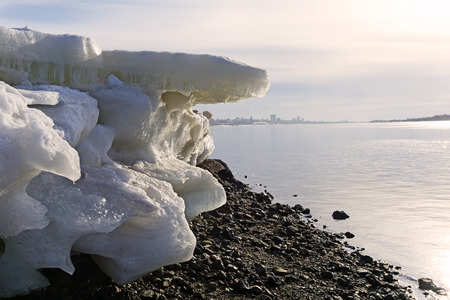 Melting ice floes on the stony beach of a wide spring river against the background of a blurred city...