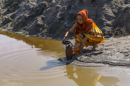 girl of oriental appearance in sari and hijab fills the pitcher with water from a dirty source in the desertの写真素材