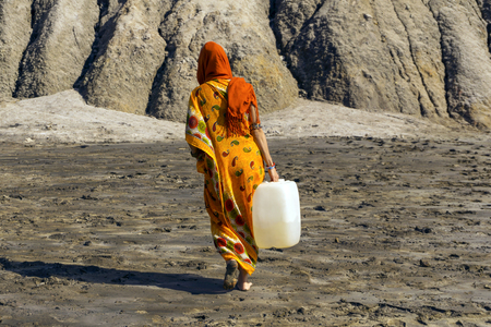 woman in a sari and hijab carries a plastic jerrican with water over a lifeless desert valleyの写真素材