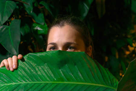girl hiding among tropical vegetation looks out from behind a wide leaf of a plantの写真素材