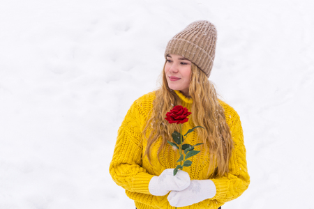 girl in a warm sweater and knitted hat holds a rose in her hands against a snowy backgroundの写真素材