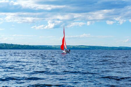 sailboat with scarlet sails performs a tacking maneuver against the background of a wooded shoreの写真素材