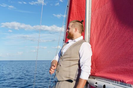 bearded man in a respectable vest looks into the distance, leaning against the mast of a sailboat in...